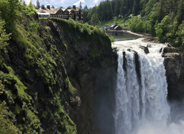 A massive waterfall surrounded by green trees and bushes. A large building is in the back left of this photo. Now the water is flowing down the fall. Everything else is still.
