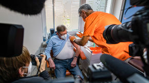Cameras point at a patient receiving a shot in the arm